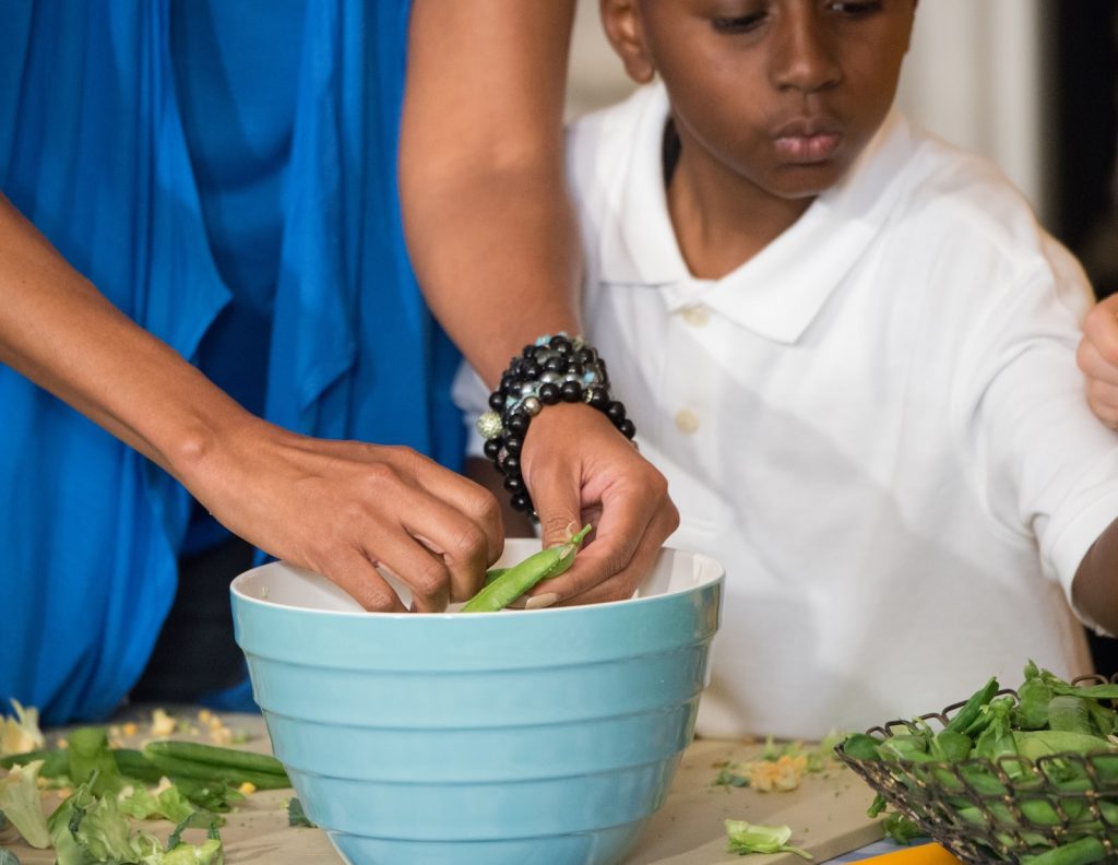 A woman’s hand putting greens into a bowl while a young boy helps.